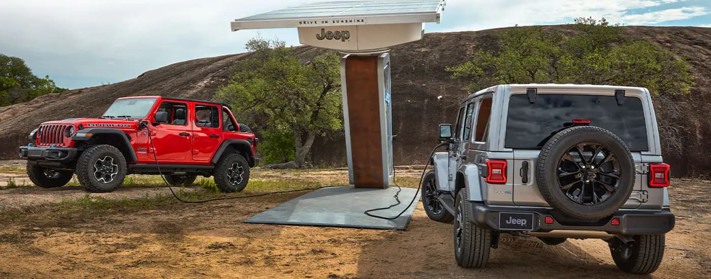 A red and a silver 2021 Jeep Wrangler 4xe are shown charging at a remote changing station.
