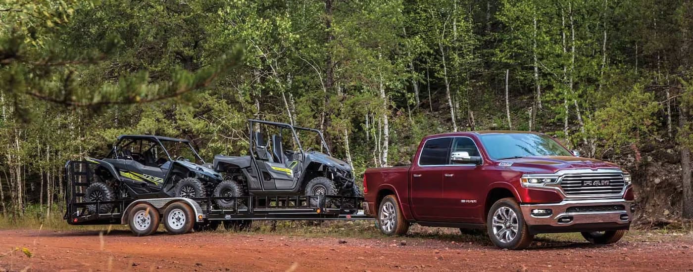 A red 2023 Ram 1500 is shown towing two UTVs on a trailer on a dirt road.